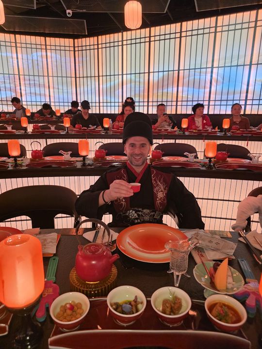 A traveller in traditional dress raising a cup at a lantern-lit banquet table